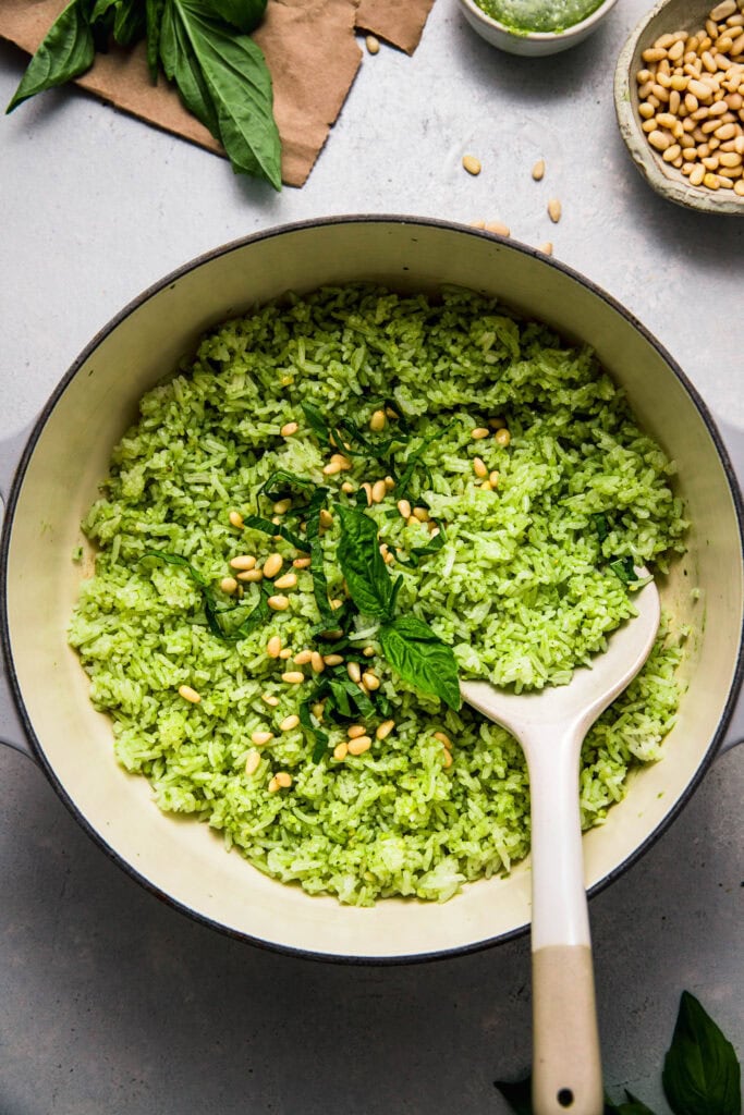 Pesto rice in pot with serving spoon, topped with basil leaves and pine nuts.