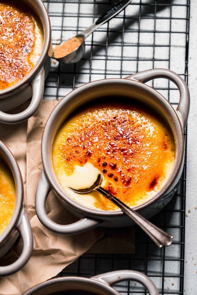 Three ramekins of creme brulee on cooling rack next to spoon of sugar.