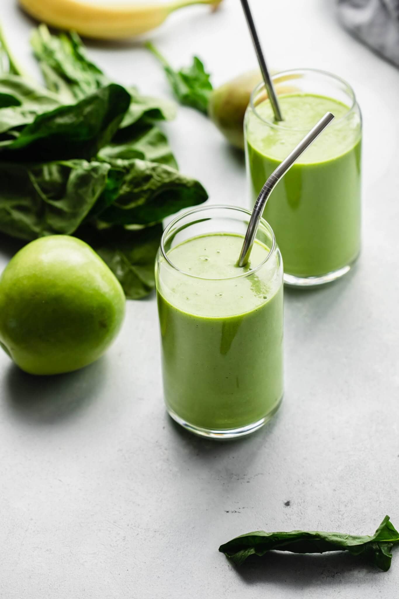 Side view of two green goddess smoothies on counter next to spinach, pear and banana.