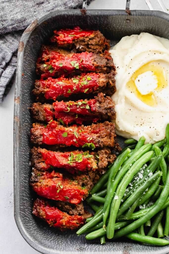Sliced meatloaf in serving tray with green beans and mashed potatoes.