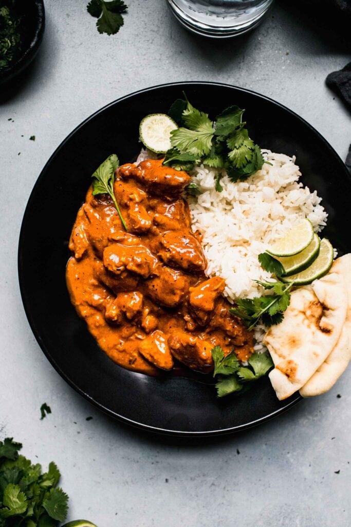 Overhead shot of indian butter chicken in black bowl with naan and lime wedges. 