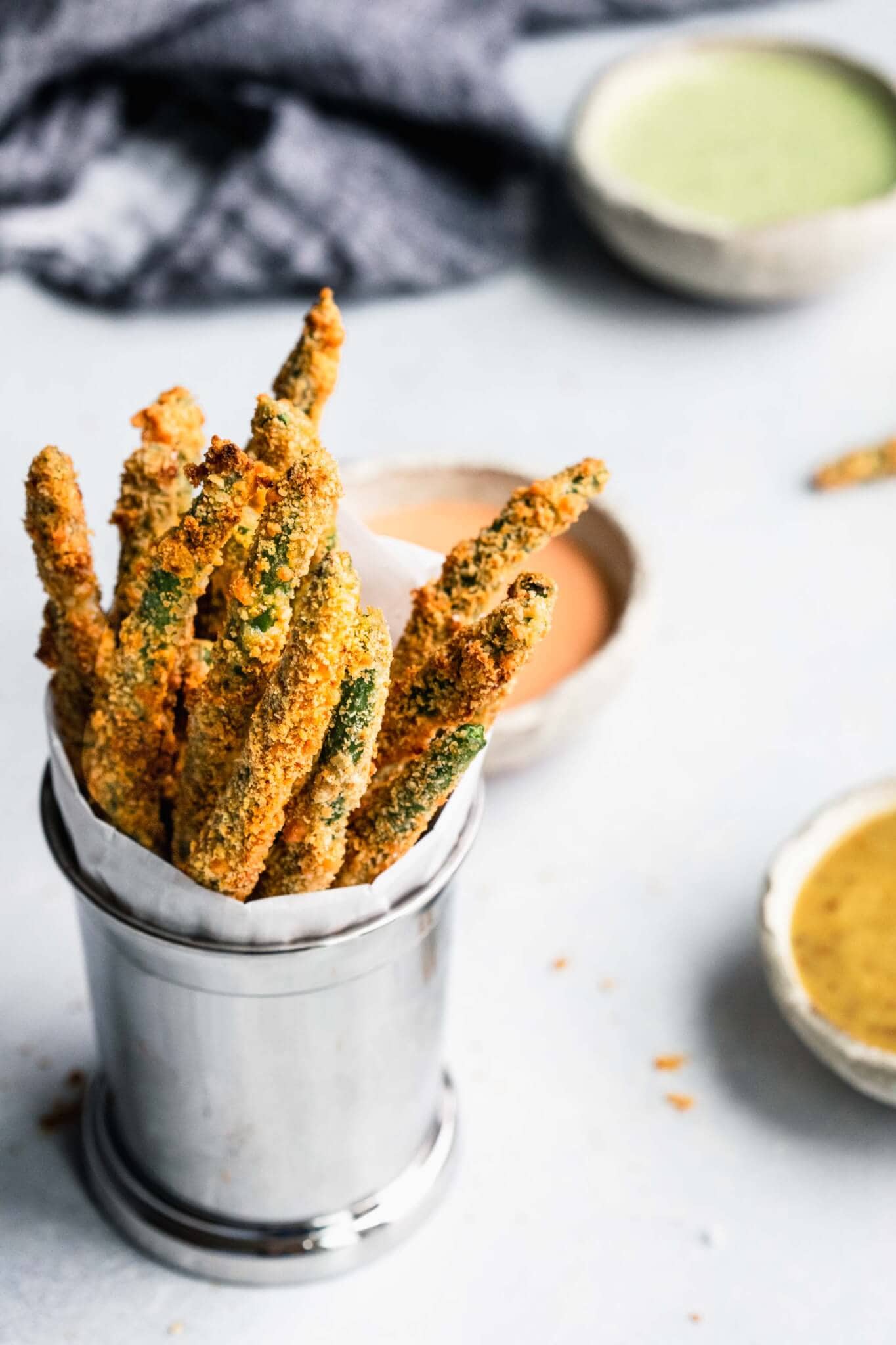 Baked green bean fries in serving cup next to three ramekins of dipping sauce. 
