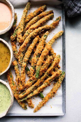 Fried green beans on baking sheet next to three types of dipping sauce.