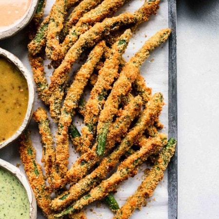 Fried green beans on baking sheet next to three types of dipping sauce.