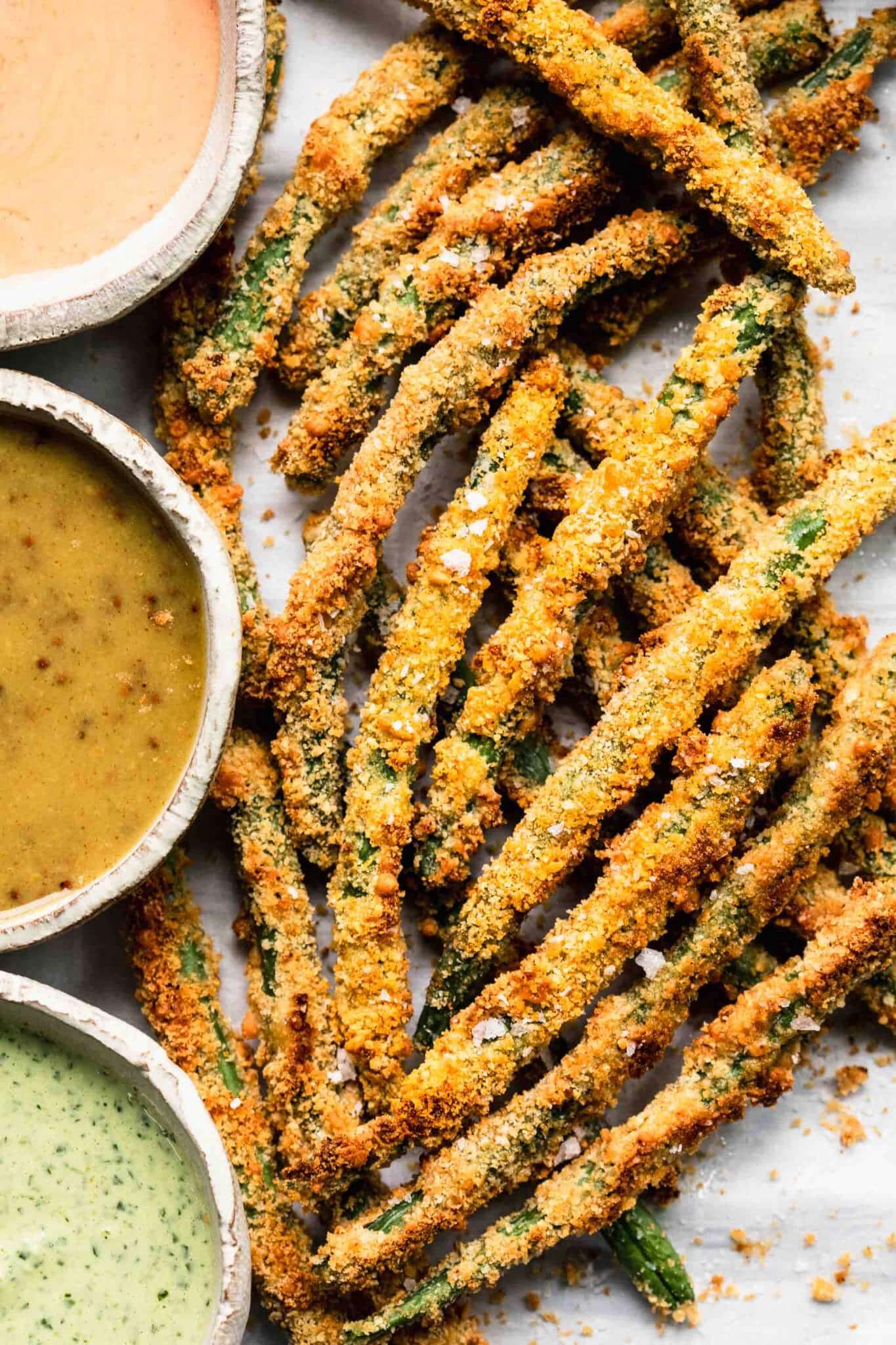 Green bean fries on baking sheet next to three types of dipping sauce.