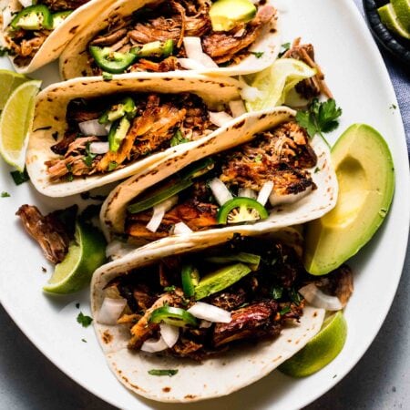 Pork street tacos arranged on white oval plate with avocado and jalapenos.