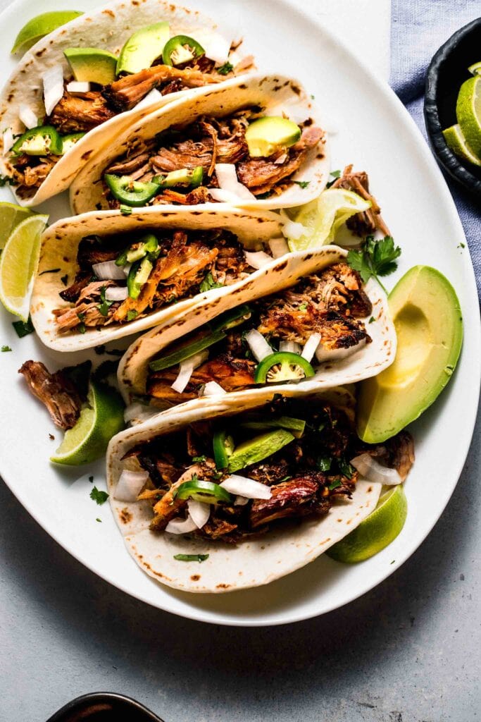 Pork street tacos arranged on white oval plate with avocado and jalapenos.