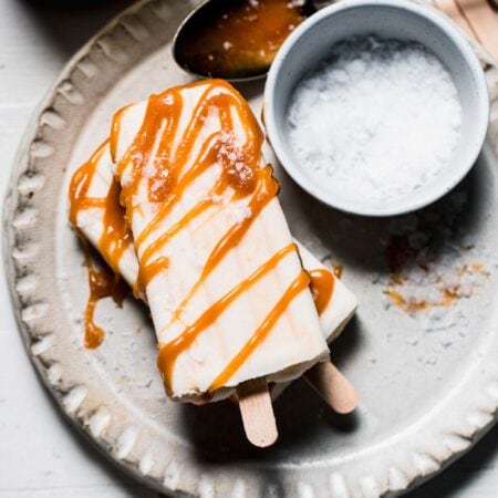 Overhead shot of two salted caramel popsicles on small plate next to bowl of salt and salted caramel in jar.