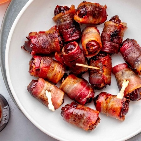 Cooked bacon wrapped dates in white serving bowl next to small bowl of almonds.