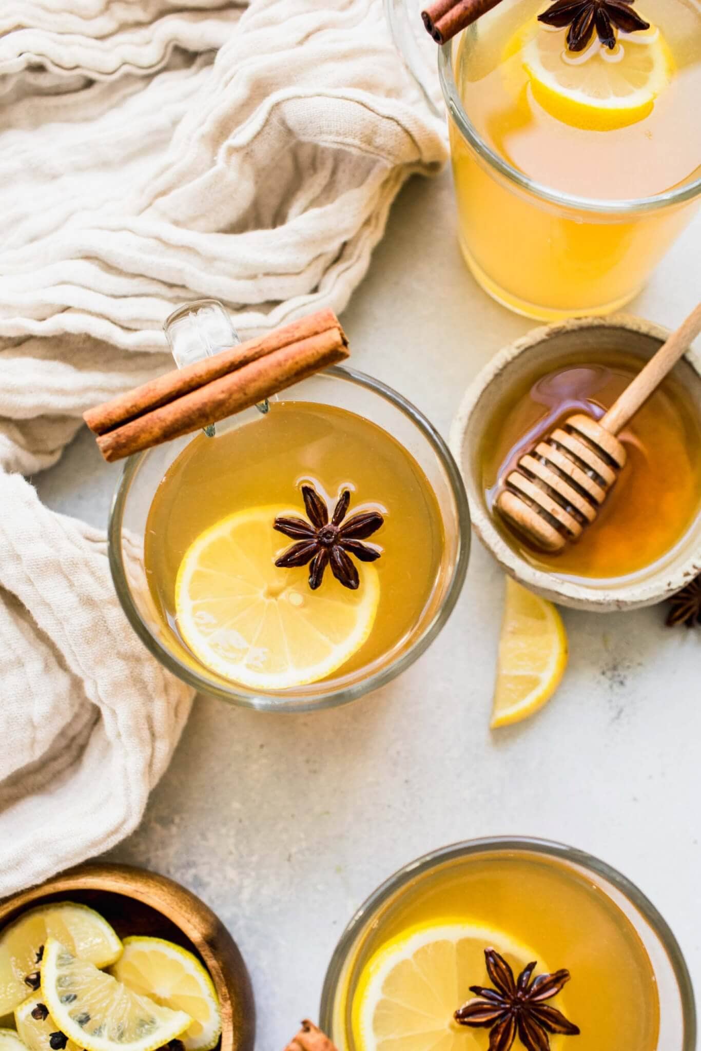 THREE MUGS OF HOT TODDIES GARNISHED WITH LEMON SLICES AND STAR ANISE NEXT TO HONEY JAR