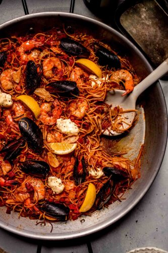 Overhead shot of fideua in paella pan topped with lemon wedges and aioli with serving spoon.