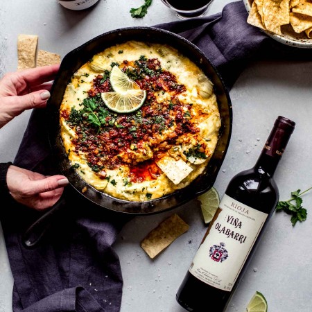 Hands holding skillet of queso blanco next to two bottles of wine.