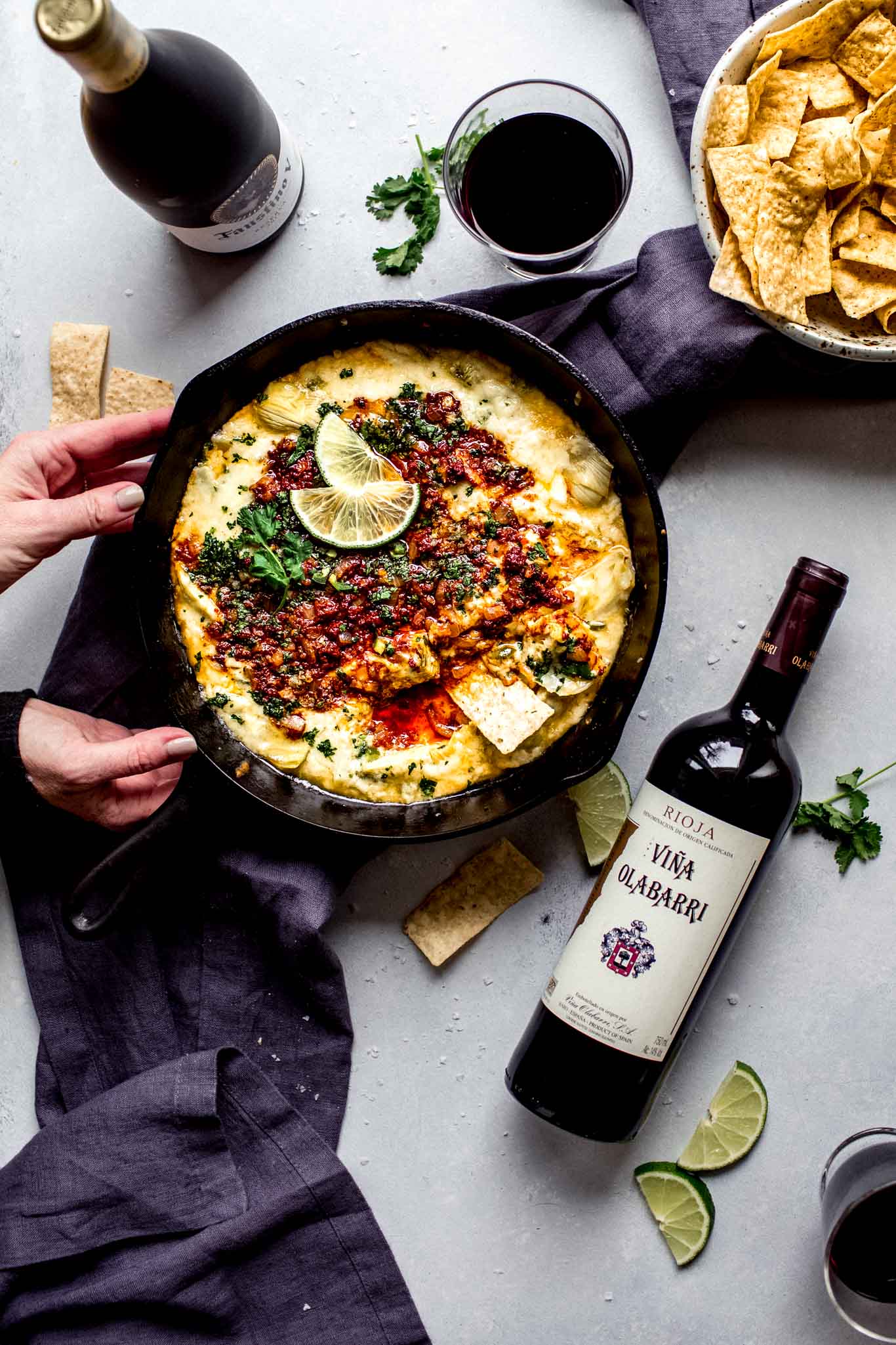 Hands holding skillet of queso blanco next to two bottles of wine.