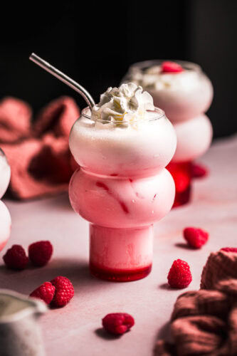 Two raspberry cream sodas on pink table with raspberries scattered around.