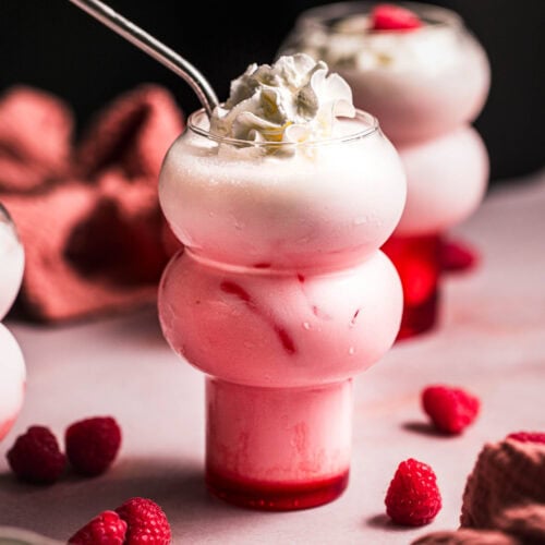 Two raspberry cream sodas on pink table with raspberries scattered around.