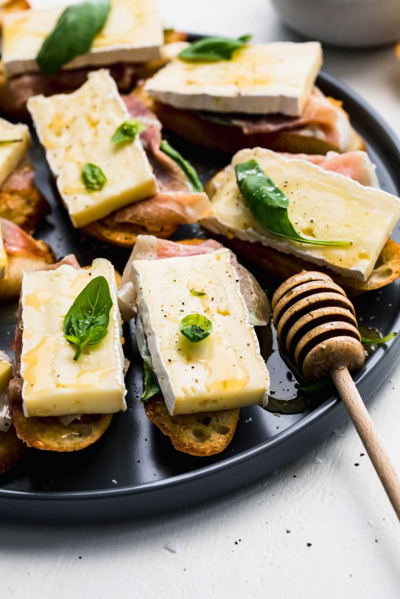 Crostini topped with brie, basil and prosciutto arranged on grey plate next to honey wand. 