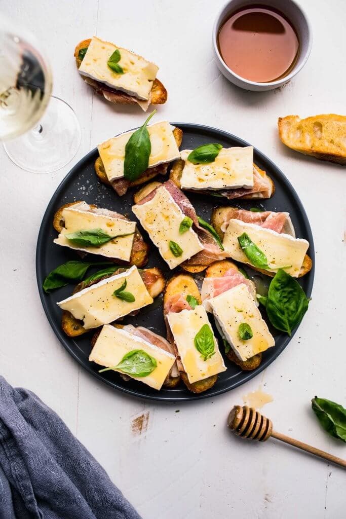 Crostini topped with brie, basil and prosciutto arranged on grey plate next to honey wand.
