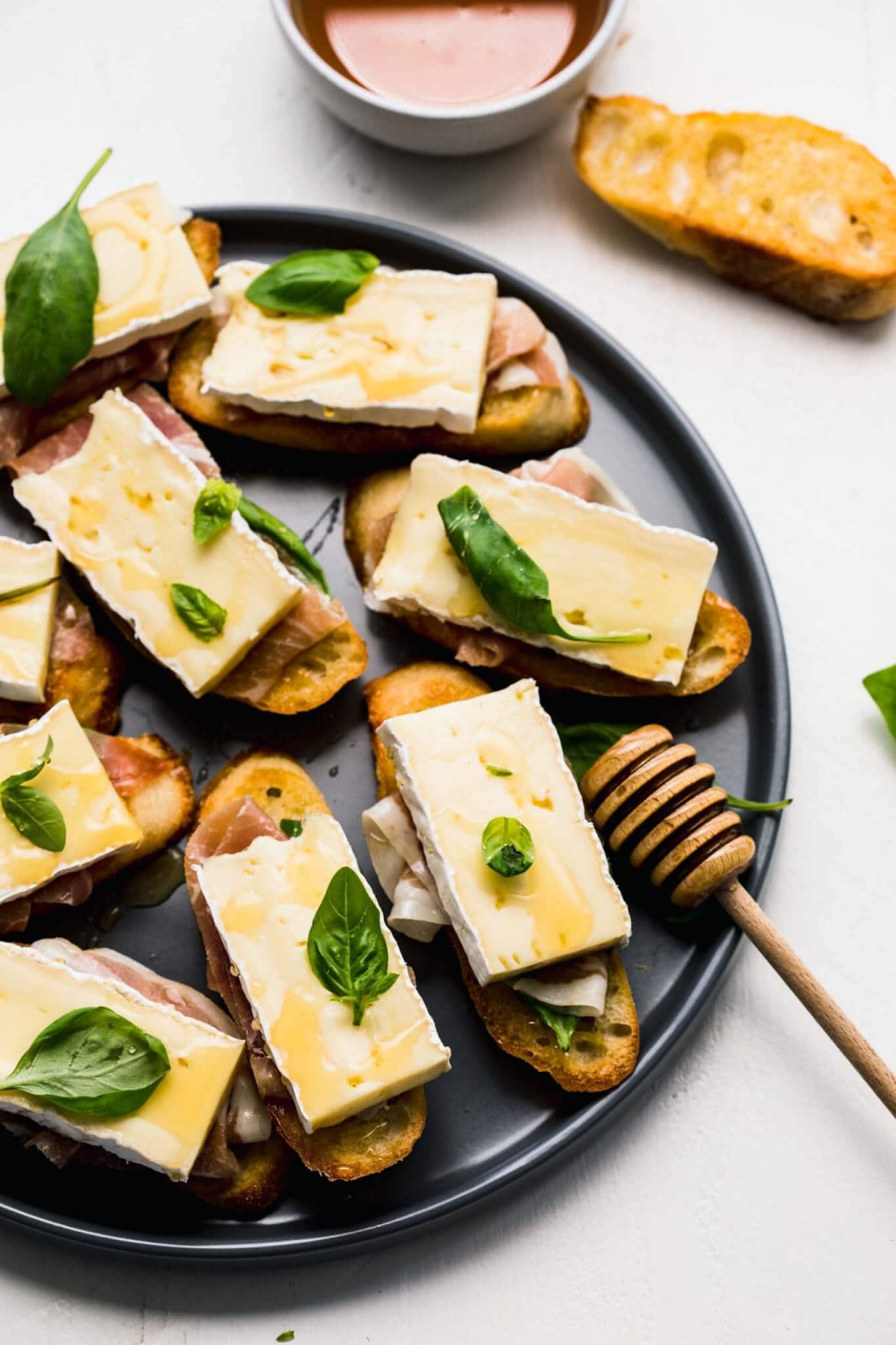 Crostini topped with brie, basil and prosciutto arranged on grey plate next to honey wand.