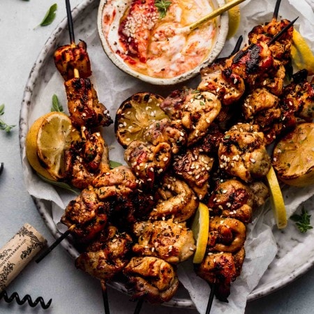 Overhead shot of lemon chicken skewers on plate next to bowl of harissa yogurt sauce.