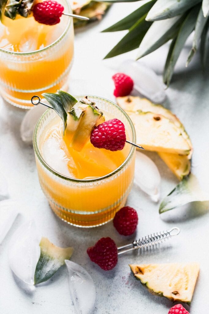 Two pineapple vodka cocktails on counter garnished with pineapple slices and raspberries.