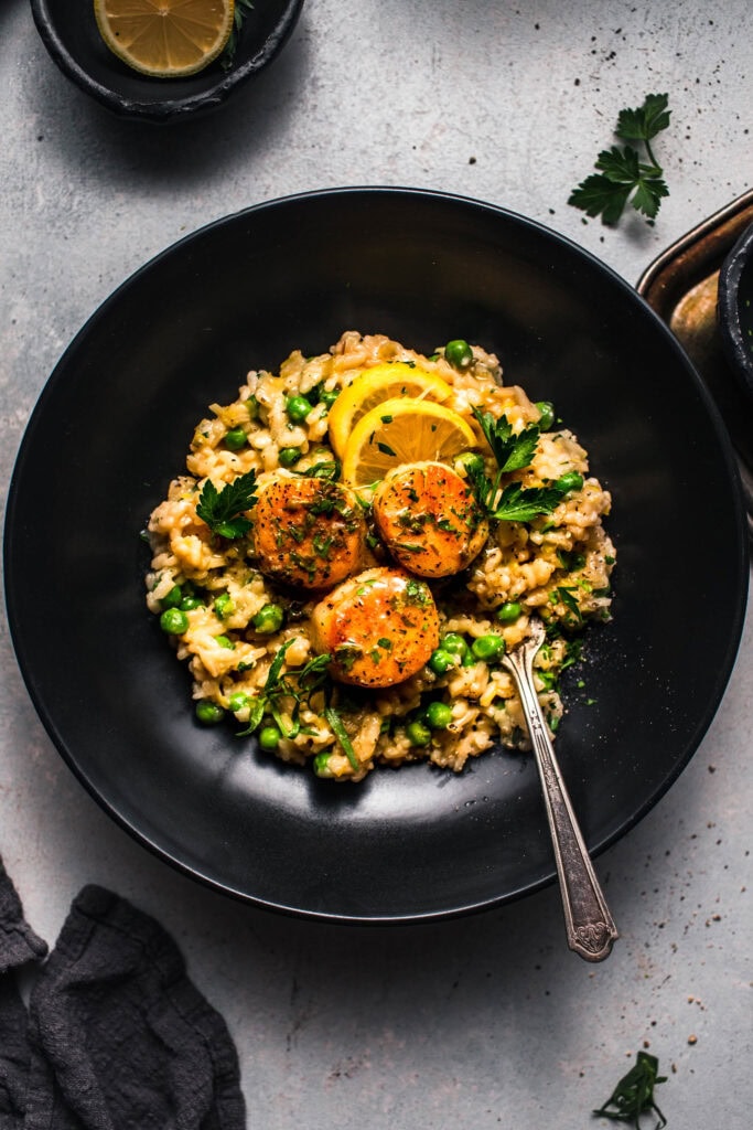 Overhead shot of pea risotto with scallops in black bowl with lemon wedges.