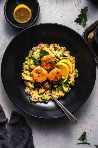 Overhead shot of pea risotto with scallops in black bowl with lemon wedges.