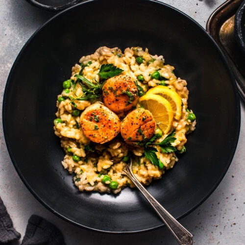 Overhead shot of pea risotto with scallops in black bowl with lemon wedges.