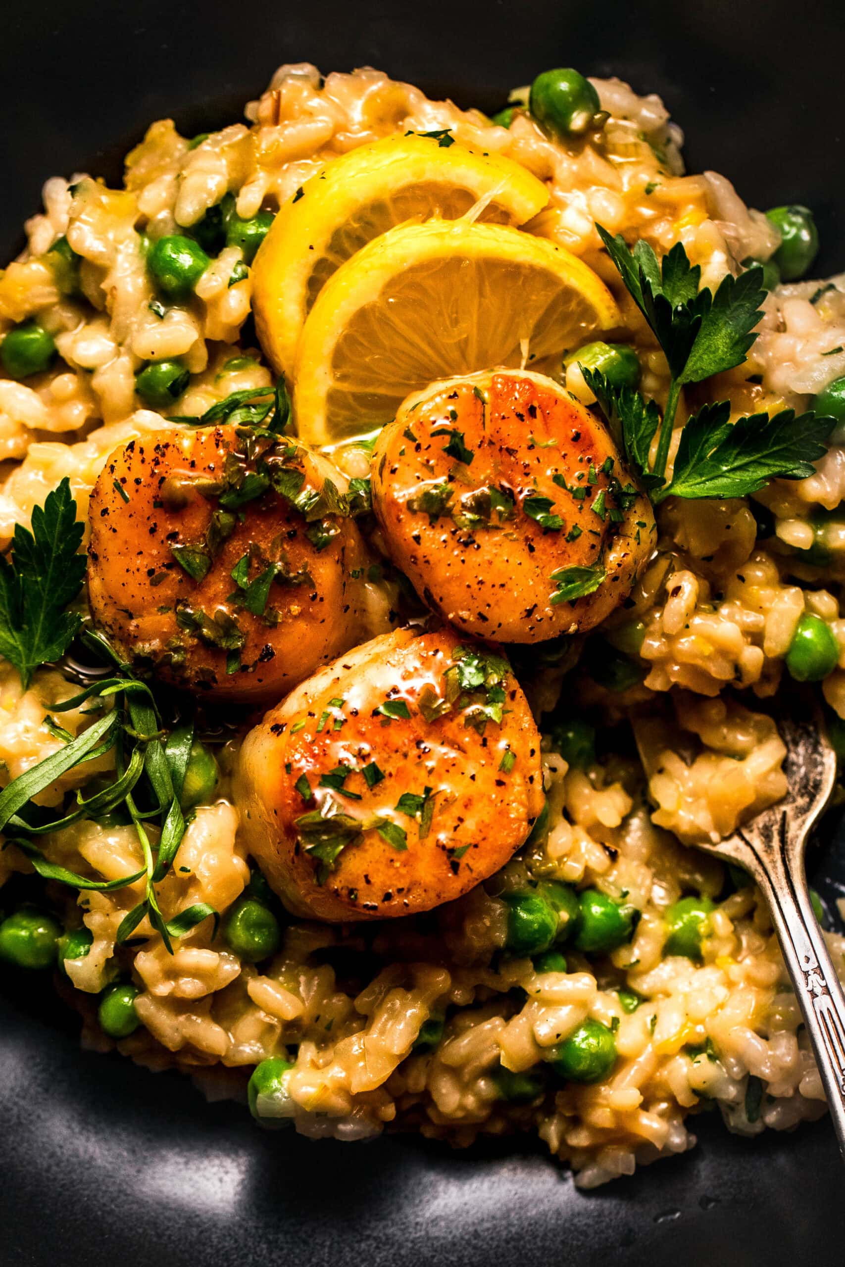 Overhead shot of scallop and pea risotto in black bowl topped with lemon wedges.