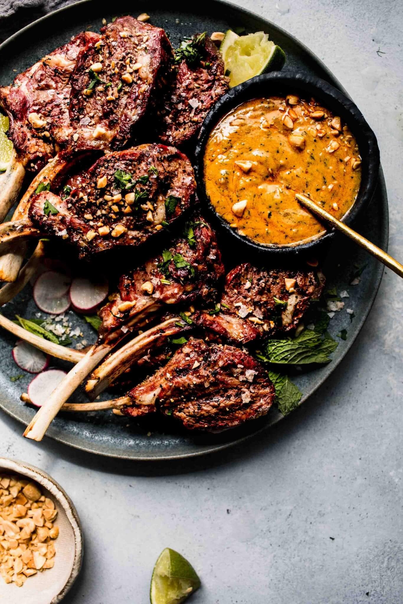 Lamb chops arranged on grey plate next to bowl of peanut sauce.