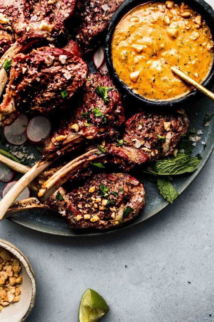 Lamb chops arranged on grey plate next to bowl of peanut sauce.