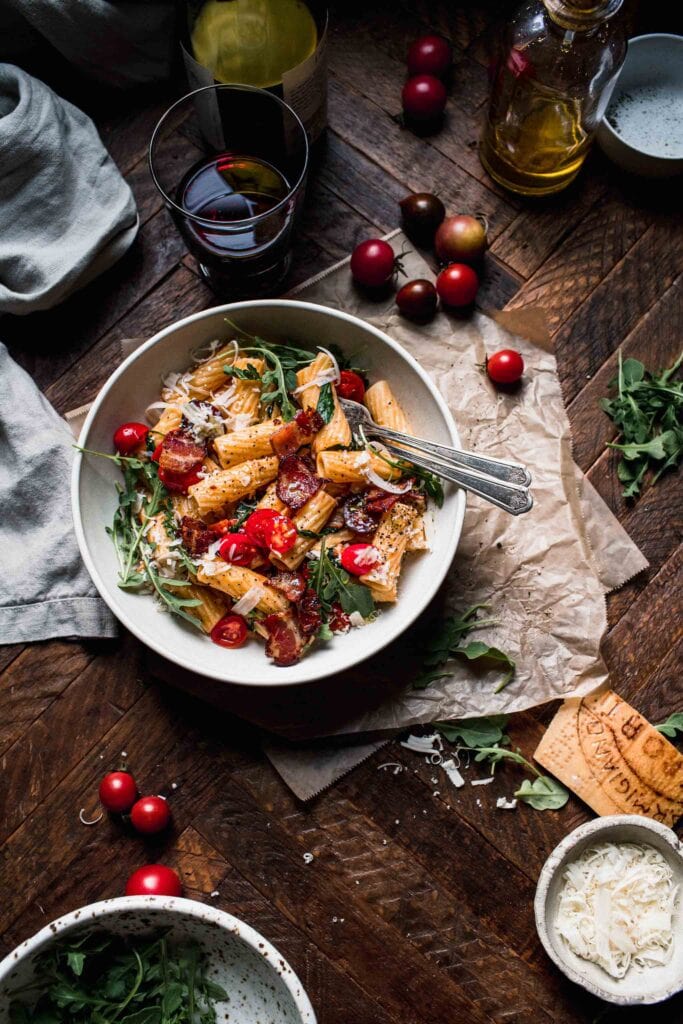 BLT pasta on wooden table next to small bowl of parmesan cheese. 