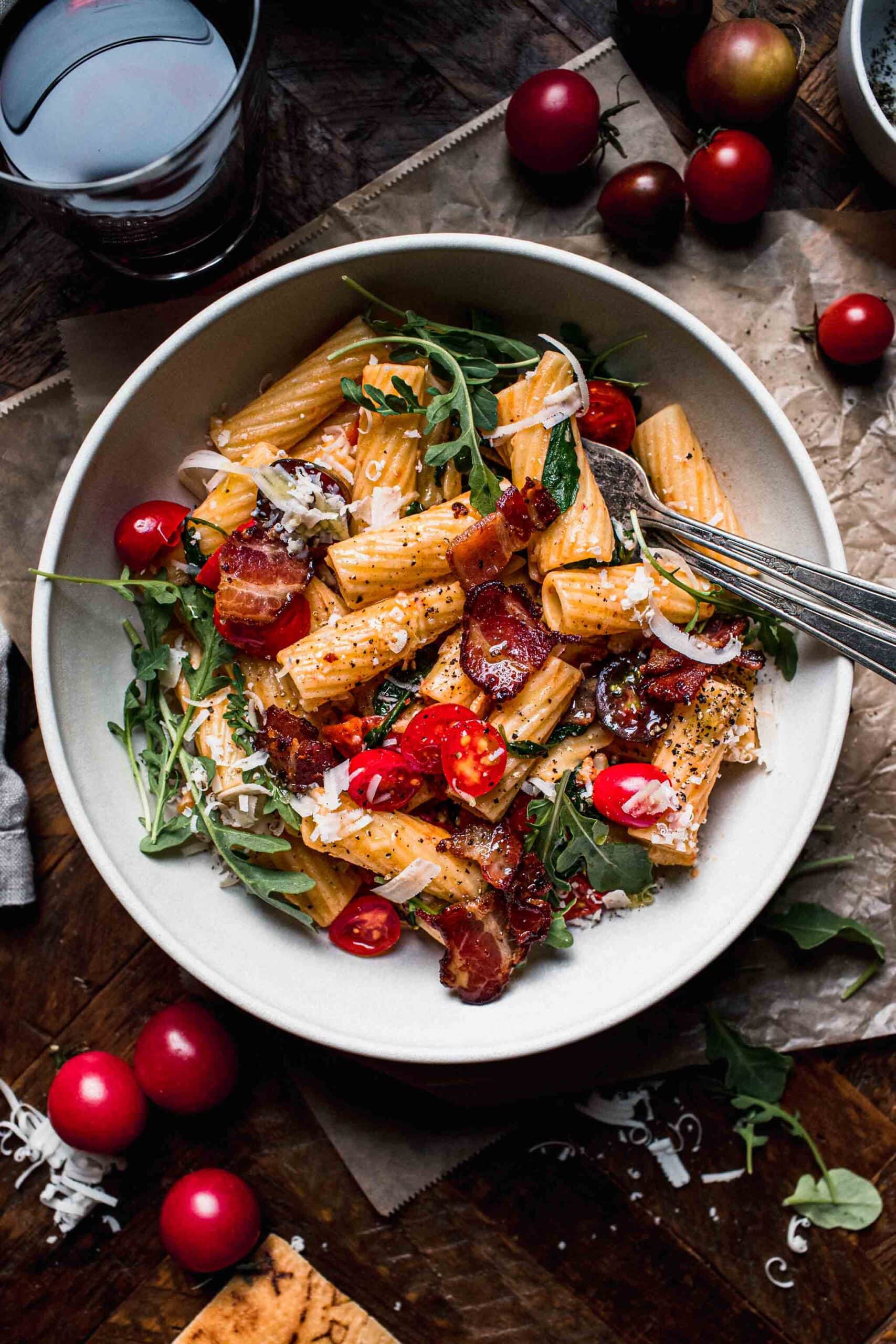 Overhead shot of blt pasta in white bowl on wood background next to glass of wine.