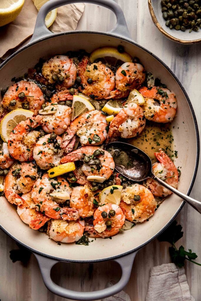 Skillet filled with shrimp piccata on pink backdrop.