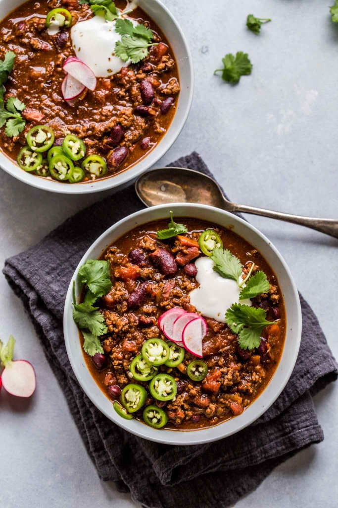 Two bowls of chili on counter ready to be eaten. topped with sour cream, radishes and jalapenos. 