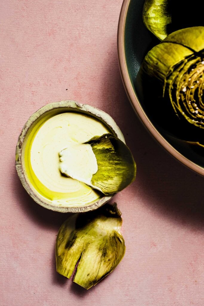 Artichoke sauce in small bowl next to large bowl of cooked artichokes. 