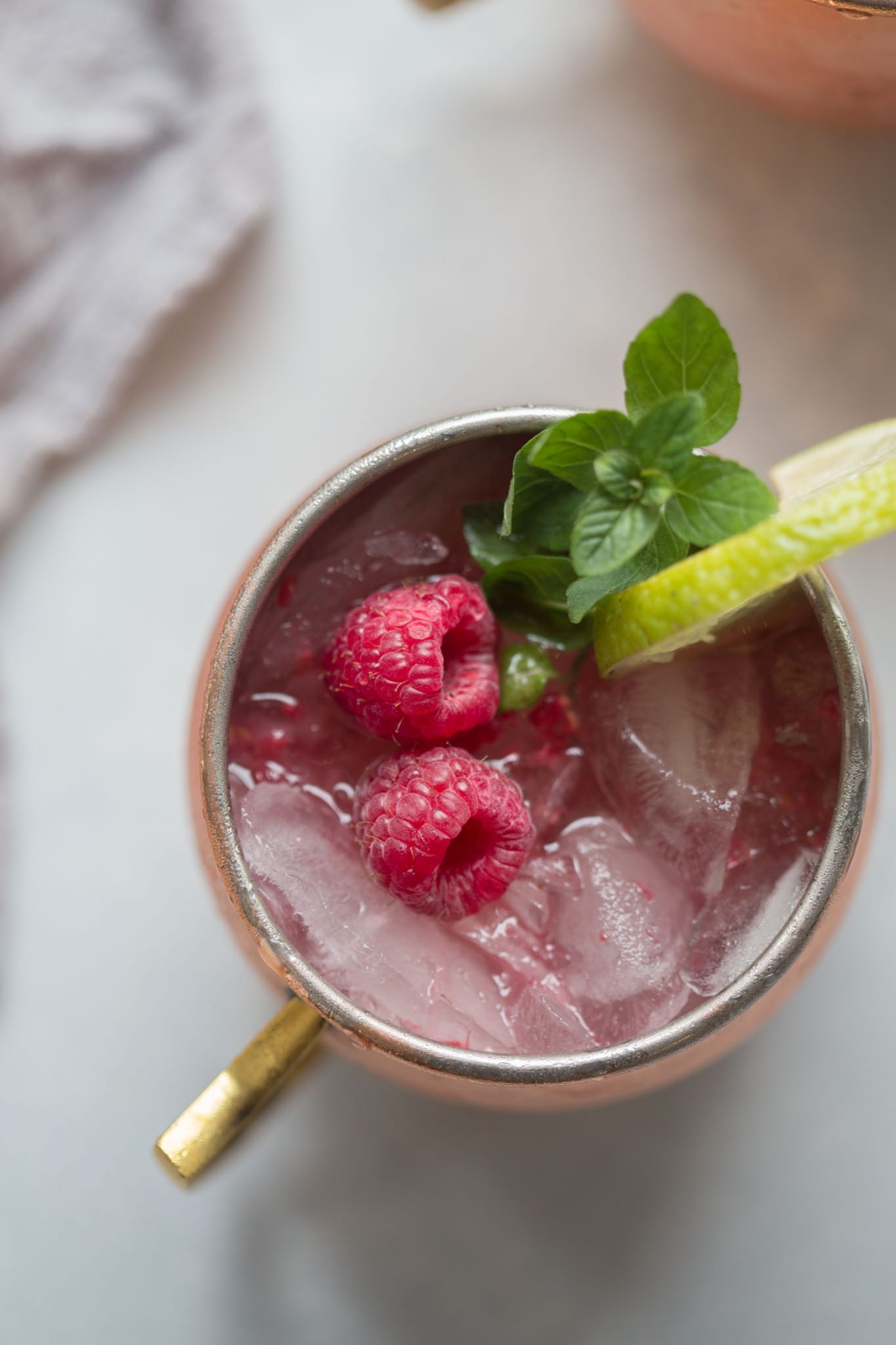 Close up mug of Raspberry Lime Moscow Mule garnished with raspberries, mint and lime in a copper mug. 