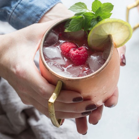 Close up of hands holding Raspberry Moscow Mule with mint leaves, lime wheel and raspberries.