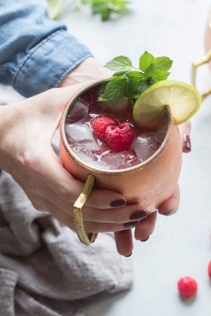 Close up of hands holding Raspberry Moscow Mule with mint leaves, lime wheel and raspberries.