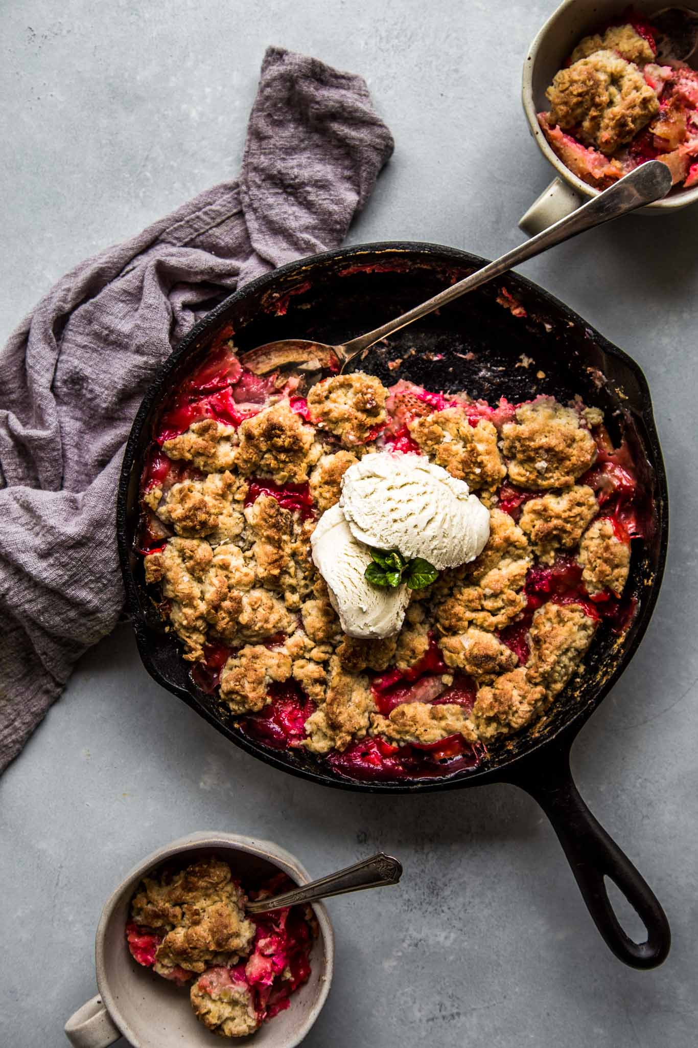 Overhead shot of cast iron skillet with strawberry rhubarb cobbler served in two mugs. 