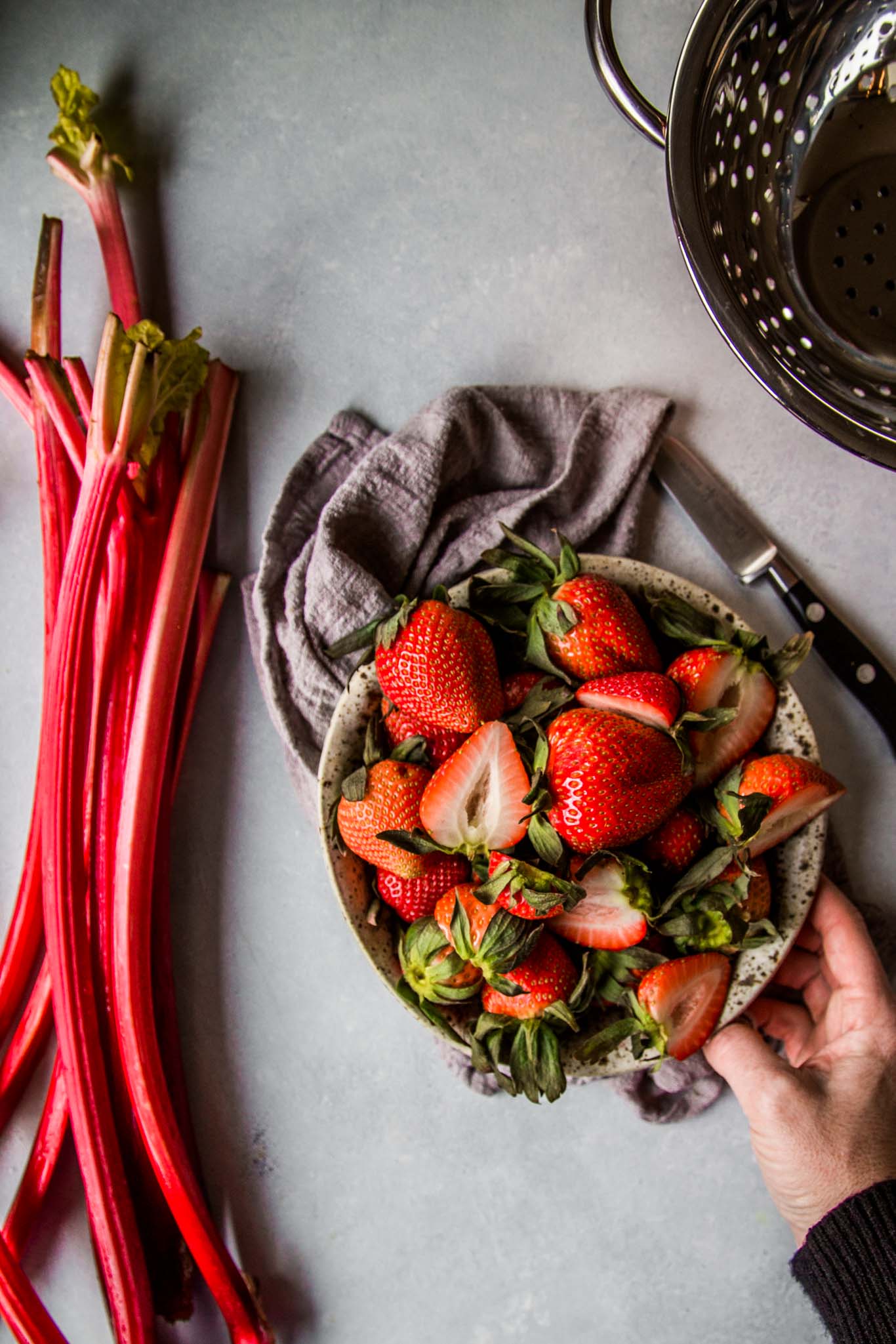 Hand reaching for bowl of strawberries next to rhubarb