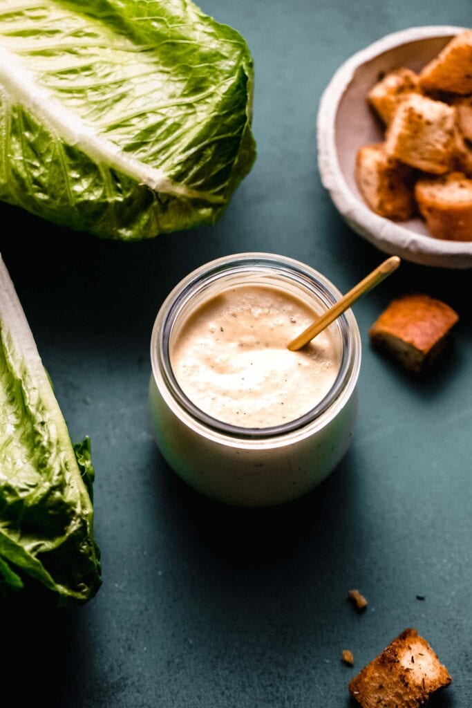 Side view of vegan caesar dressing in small jar with spoon. 