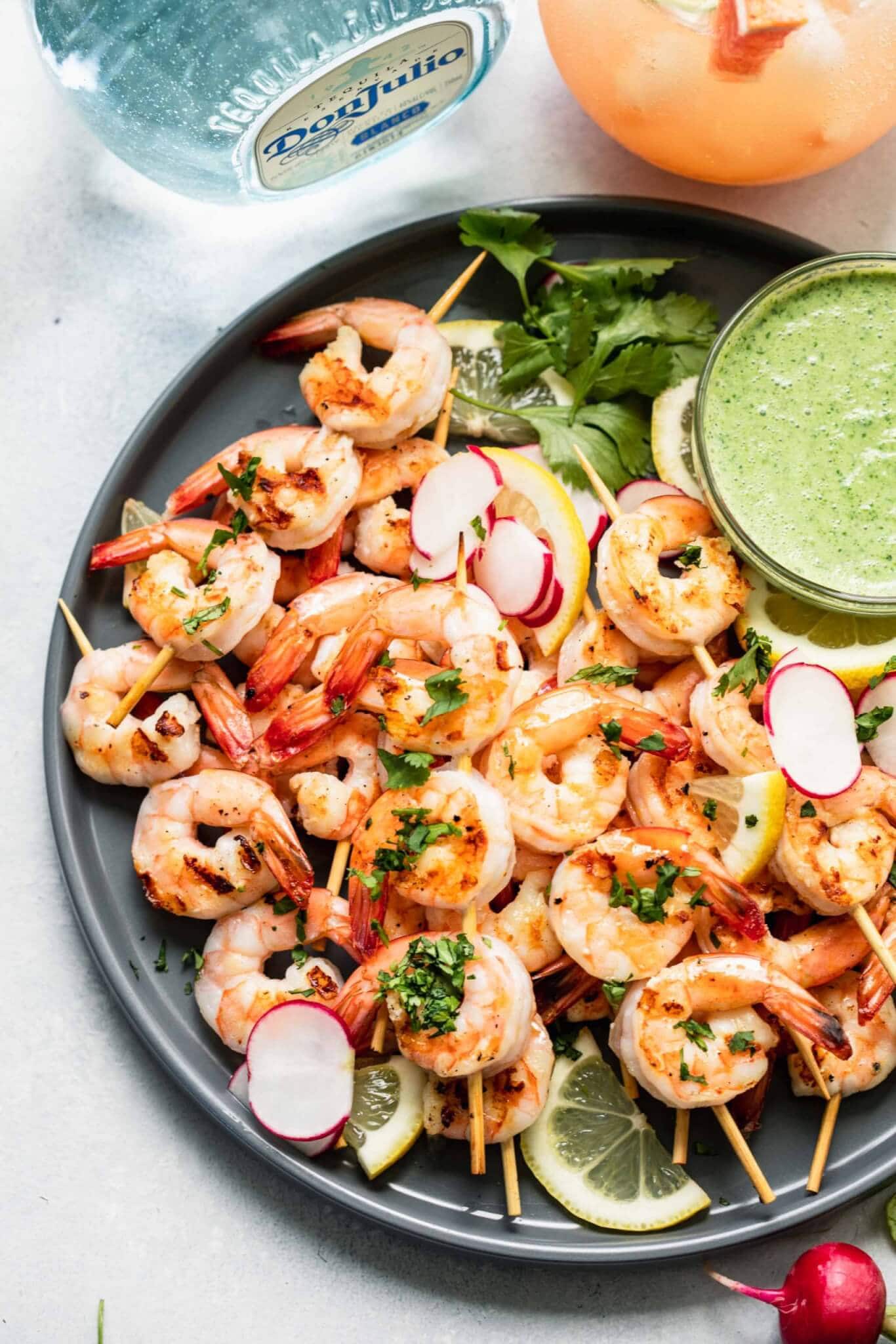 Cooked shrimp skewers arranged on a grey plate with bowl of creamy green sauce next to bottle of tequila and paloma cocktail.