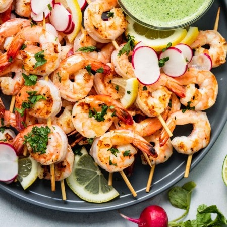 Cooked shrimp skewers arranged on a grey plate with bowl of creamy green sauce.