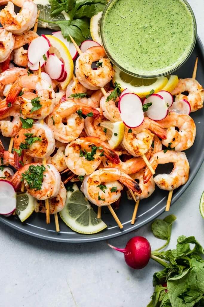 Cooked shrimp skewers arranged on a grey plate with bowl of creamy green sauce.