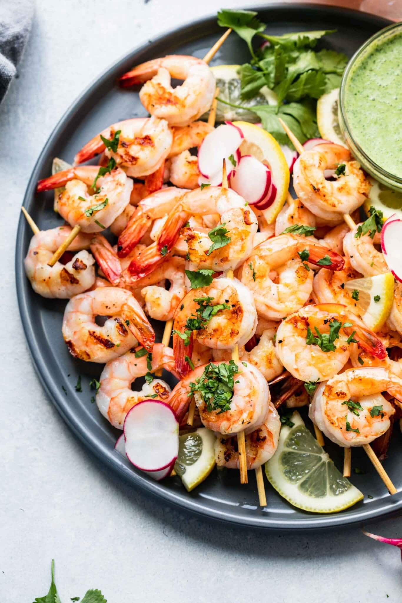 Cooked shrimp skewers arranged on a grey plate with bowl of creamy green sauce.