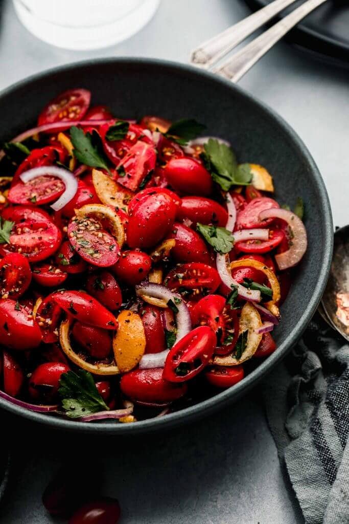 SIDE VIEW OF CHERRY TOMATO SALAD IN GREY BOWL WITH SERVING SPOONS.