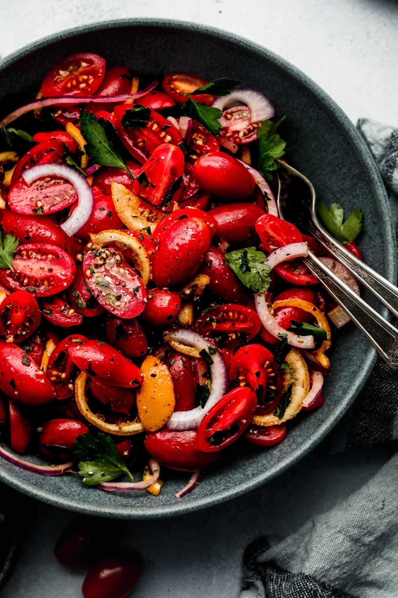 SIDE VIEW OF CHERRY TOMATO SALAD IN GREY BOWL WITH SERVING SPOONS.