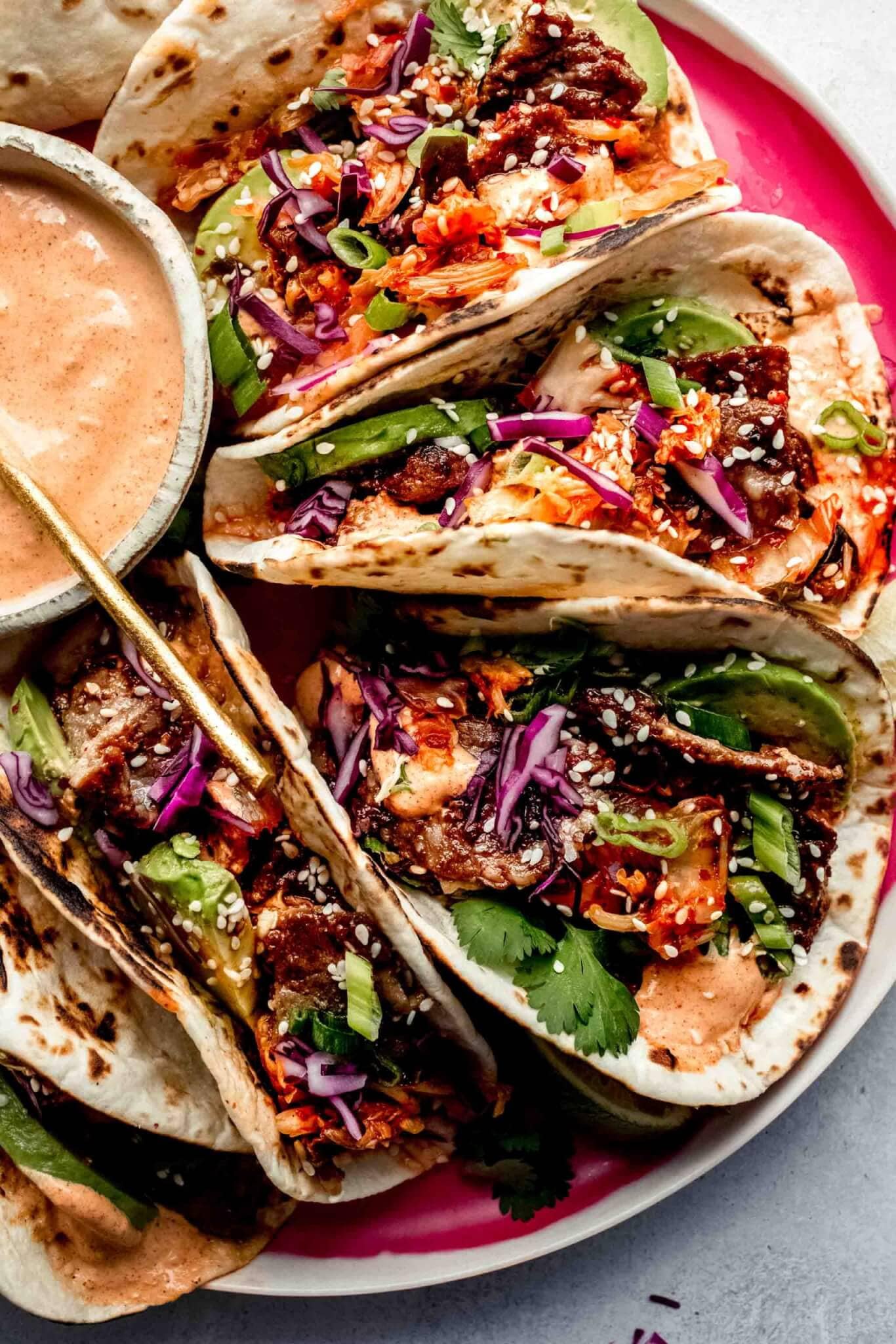 Korean tacos arranged on bright pink plate next to small bowls of kimchi and aioli.