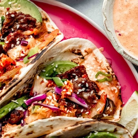 Korean tacos arranged on bright pink plate next to small bowls of kimchi and aioli.