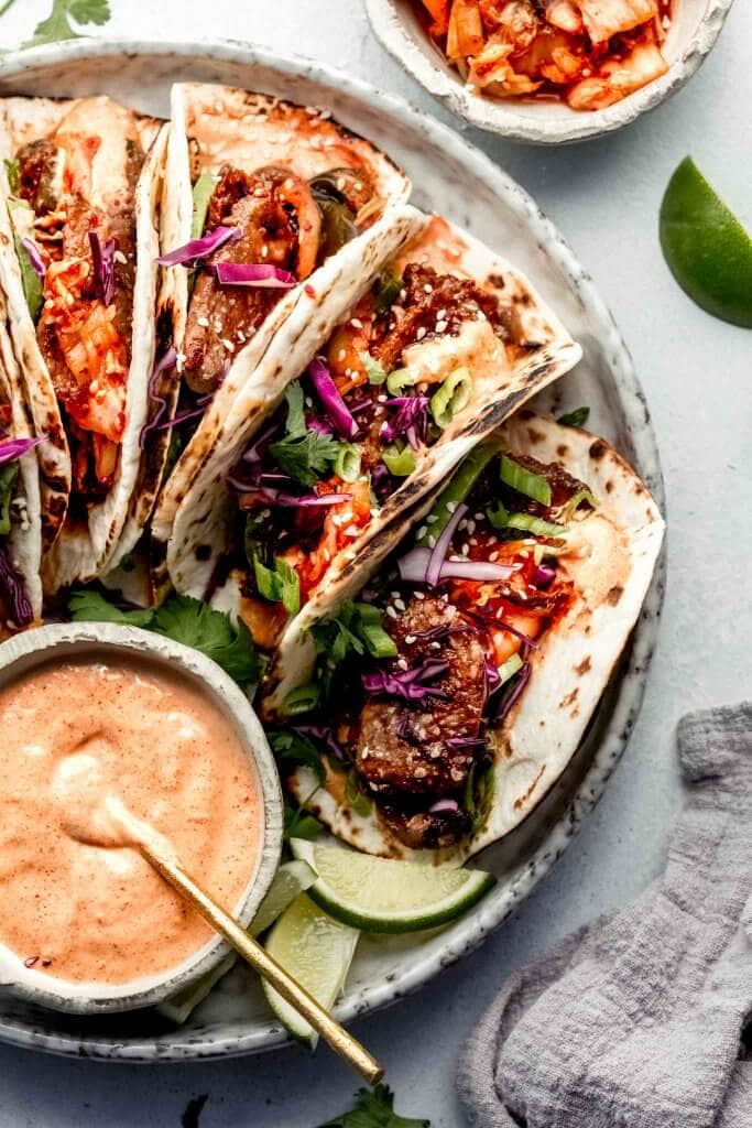 Kimchi tacos next to bowl of aioli on grey plate. 